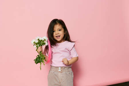 a cute little preschool girl stands on a pink background in a pink T-shirt and shows a bouquet of white daisies, actively moving her hands, smiling happily. The theme of childrens delightの写真素材