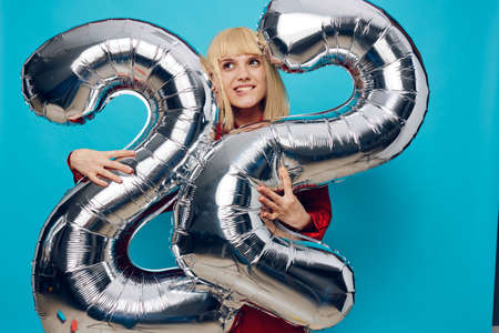 a woman in a red shirt stands showing her teeth on a blue background and holds inflatable balloons in the shape of the number twenty-two in silver color hugging them with her handsの写真素材
