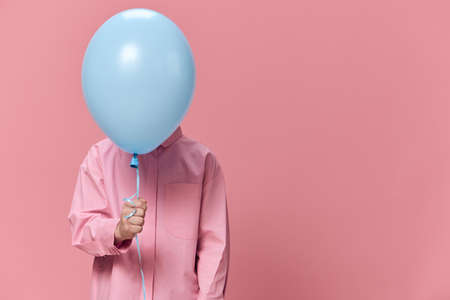 a cute, funny boy of school age stands on a pink background in pink clothes and funny hides his face...