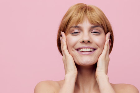 photo with an empty space of a pleasant, lovely woman with perfect skin, standing on a pink background, gently touching her face with her hands, squinting her eyes with pleasure and smiling prettily.の写真素材