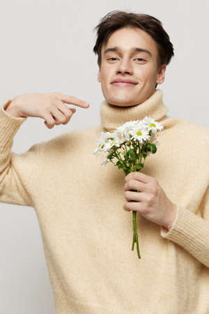 Vertical studio shot. close-up photo.An emotional young man with dark, short, beautiful hair combed back in a beige turtleneck with a high neck stands on a gray background with a bouquet of daisiesの写真素材