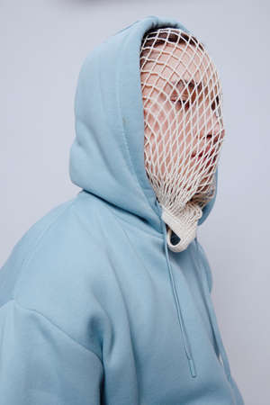 a man suffering from problems with a mesh on his face is standing in a light blue hoodie on a light...