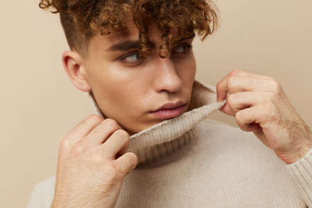 a handsome, attractive man stands on a beige background in a light turtleneck and stretches the neck with his hands. Horizontal photoの写真素材