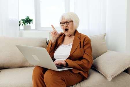 an angry, aggressive elderly businesswoman is holding a video conference through a laptop, holding it on her lap and shouting loudly showing her index finger upの写真素材