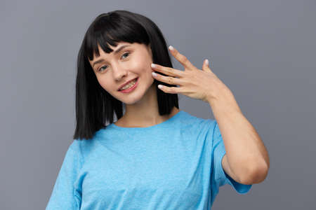an attractive sweet woman stands on a gray background in a blue T-shirt touches her face with her fingertips smiling in surpriseの写真素材