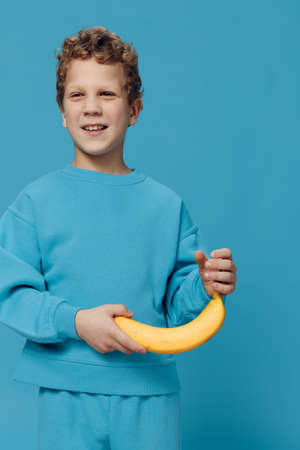 a beautiful, happy boy stands in blue clothes on a blue background and holds a banana in his hand smiling joyfully. Vertical studio photoの写真素材