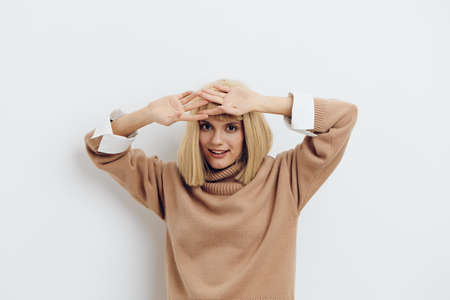 Horizontal photo, a woman on a white background in a beige sweater with beautiful blond hair looks at the camera with her hand raised above her headの写真素材