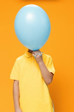 a boy of school age stands on an orange background in a yellow polo and covers his face with a large blue balloonの写真素材