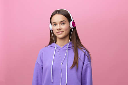 a nice, sweet, attractive woman is standing in a purple tracksuit listening to music in her headphones and looking sweetly into the camera standing on a pink background with an empty spaceの写真素材