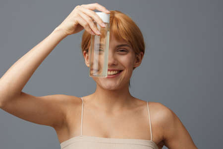 Horizontal studio shot. A nice woman in a pale white T-shirt with illuminating clear skin, with ginger silky hair gathered in a ponytail on a gray background with a transparent bottle of water.の写真素材