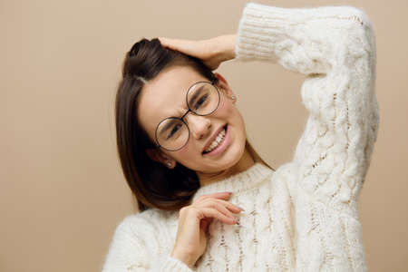 a pretty woman stands on a light background in a white sweater and glasses, straightens her hair with one hand, throwing it back, frowning a littleの写真素材
