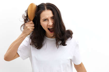 sad, upset woman screams in pain trying to comb her long, black hair with a massage comb, looking at the cameraの写真素材