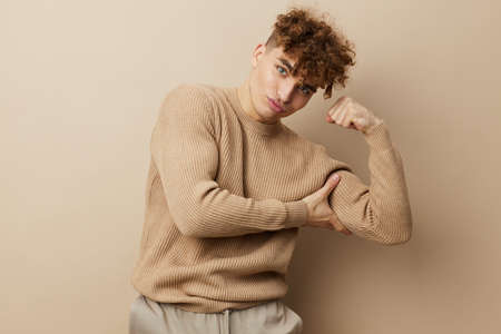 a charming, handsome man stands on a beige background in a sweatshirt and shows his biceps muscles with his handの写真素材