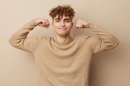 a charming, handsome man stands on a beige background in a sweatshirt and shows the muscles on his armsの写真素材