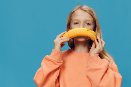 handsome, happy girl stands in orange clothes on a blue background and holds a banana in her hand, substituting it as a smile to her face. Studio photo with empty space for advertising insertの写真素材
