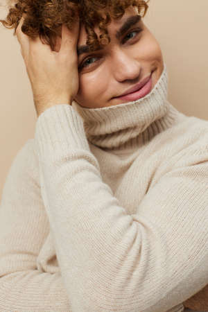 a handsome, attractive man with curly hair stands on a beige background in a light turtleneck pulled over his face, looking pleasantly into the camera, holding his hand on his head in his hairの写真素材