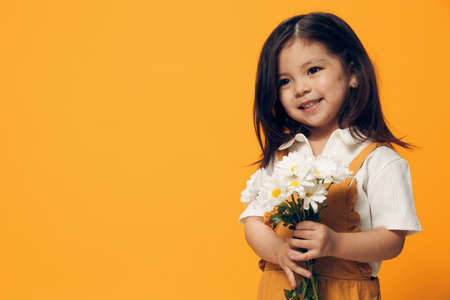 an adorable, attractive little girl in an orange sundress stands with a bouquet of daisies in her hand and smiles joyfully looking at the camera.の写真素材