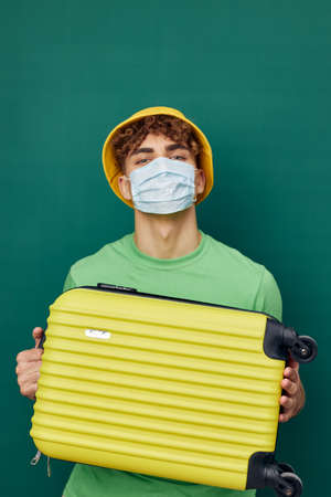 a handsome man stands on a green background, dressed in a yellow panama hat and with a mask on his face, holding a bright travel suitcase in his hand and looking at the cameraの写真素材