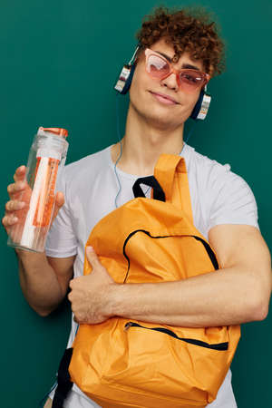 a man in gray clothes stands listening to music in headphones with bright glasses on his face, and...