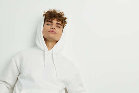 a cute man stands on a white background in a white hoodie with a hood on his head and looks...