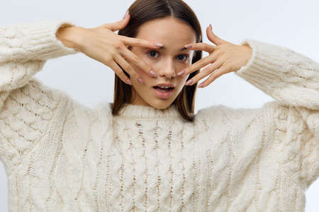 a close horizontal portrait of a beautiful, sophisticated woman standing on a light background in a knitted sweater and touching her face with a gentle movement of her fingers.の写真素材