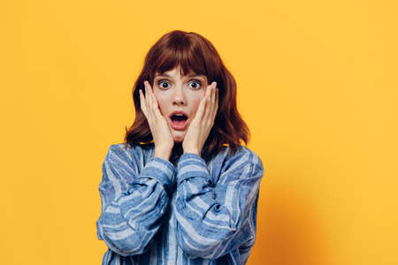 a surprised, sweet woman stands on a yellow background in a blue shirt and holds her hands near her...