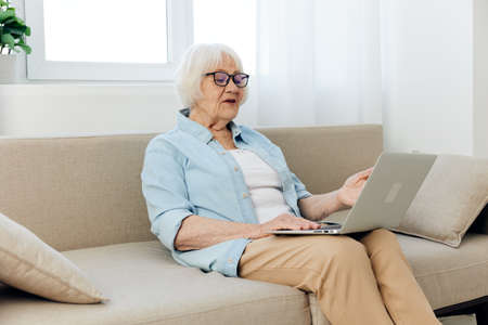 a sweet, joyful pensioner in a light shirt is sitting in a beautiful interior on a beige sofa near...