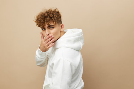 a happy, attractive man stands on a beige background in a light hoodie with his back to the camera, holding his hand near his lips, and turning his head backの写真素材