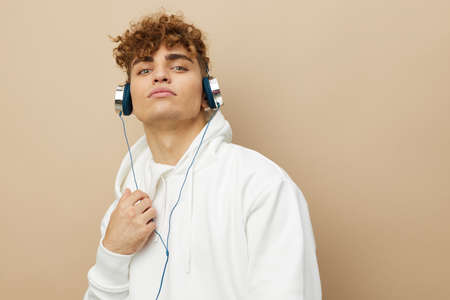 a cute, attractive man stands on a beige background in a white hoodie and listens to music with headphones, holding the wire with his hand. Horizontal photoの写真素材