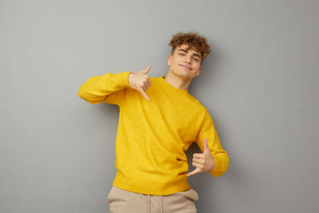 man in a yellow sweater stands on a gray background and happily looking at the camera shows gestures with his hands, standing with his back against the wall. Photo for inserting an advertising layoutの写真素材