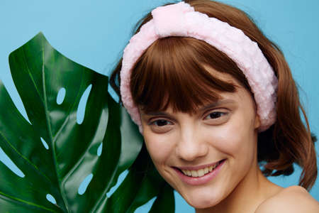 photo of a beautiful, young woman with clear skin with a soft rim on her head standing on a blue background holding a large leaf of a tropical plant in her hand and smiling pleasantlyの写真素材
