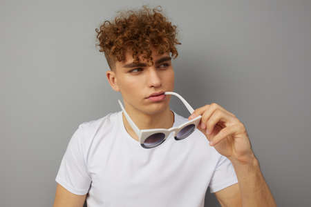 a young man stands in a casual T-shirt on an isolated gray background holding white glasses in his hands playfully biting their frameの写真素材