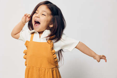 a cute little girl of preschool age is standing in an orange dress on a white background with her hands on her belt in fright and looks at the cameraの写真素材