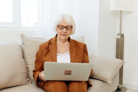 an attentive, pleasant elderly woman is sitting in a bright room at home on a cozy sofa and is...