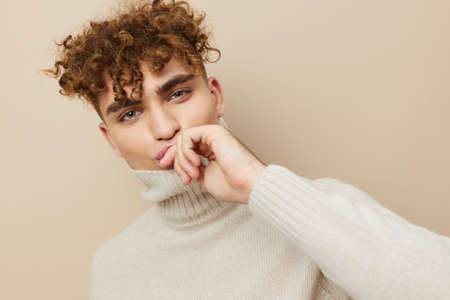 a handsome, attractive man with curly hair stands on a beige background in a light turtleneck pulled...