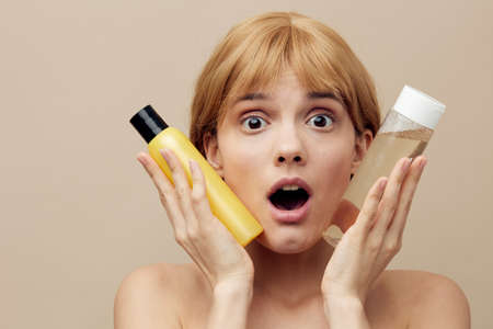 a happy, shocked young woman with clean skin stands on a beige background and holds jars of cosmetics in her hands, bringing them close to her face, her mouth wide open in surpriseの写真素材
