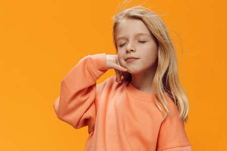 sweet girl, blonde, of school age, stands on an orange background in bright clothes and shows a...