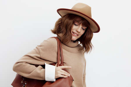 a happy woman stands on a white background in a sweater, shirt and hat holding a bag on one shoulder...