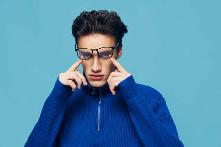 a handsome, attractive man stands on a light blue background in a blue zip-up sweater and holds his fingers touching his eyes behind glasses on his faceの写真素材