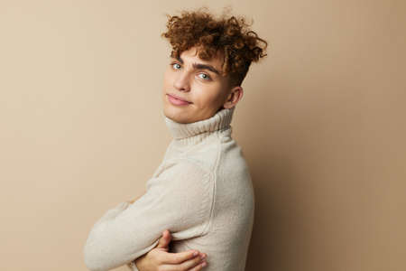 portrait of a handsome man with curly hair, standing on a beige background in a light turtleneck,...