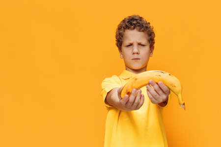 a cute, curly-haired boy with a sad face stands in yellow clothes on a yellow background and holds out a banana to the cameraの写真素材
