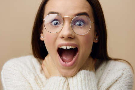 an emotional, funny, playful woman stands with her mouth wide open in surprise on a beige background in a white sweater and round eyeglassesの写真素材