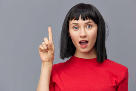 a surprised, funny woman stands on a gray background in a red T-shirt and points her index finger...