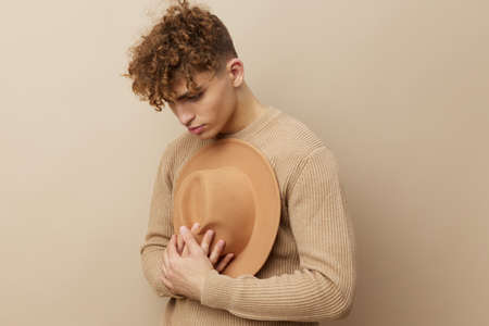 a handsome, young man stands on a beige background in a stylish sweater and holds a wide-brimmed hat in his hand, clutching it to his chest.の写真素材