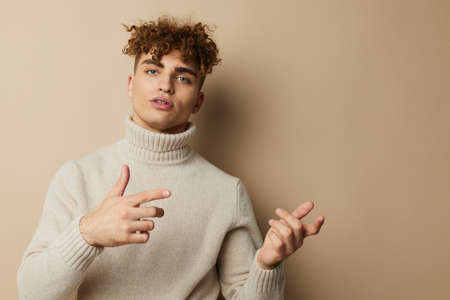 portrait of a handsome man, with curly hair, standing on a beige background in a light turtleneck,...