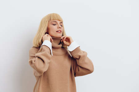 Horizontal photo, a woman on a white background in a beige sweater with beautiful blond hair closed her eyes and straightens the collar of her sweaterの写真素材