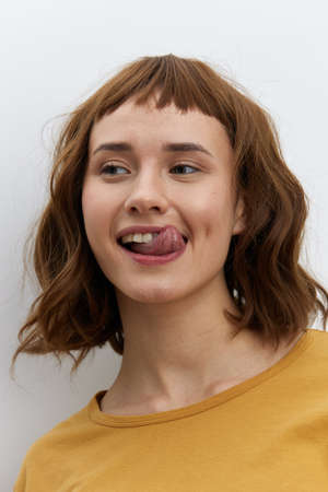 horizontal close-up portrait of a beautiful, playful, tanned woman in a yellow T-shirt on a light background amusingly showing her tongue to the camera smiling broadlyの写真素材