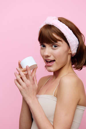 a happy, joyful, young woman stands with a soft pink rim on her head holding a white jar of cream. Vertical photographyの写真素材