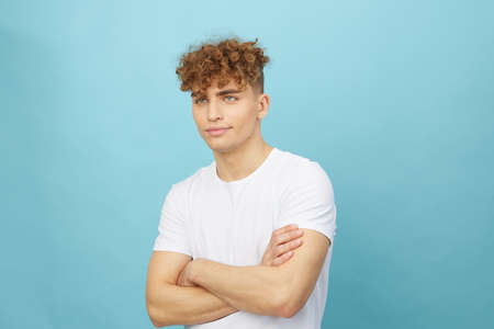 an attractive man with curly hair stands on a light blue background in a white cotton T-shirt and looks at the camera with his arms crossed on his chest. Horizontal studio photography with empty spaceの写真素材