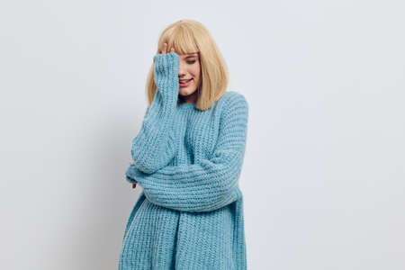 a happy, smiling woman stands on a light background in a blue sweater and holds her hand covering her faceの写真素材